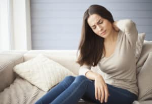 brunette girl sitting on the couch at home with a headache and back pain.